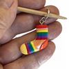One large rainbow coloured resin sock charm stitch marker on bamboo needle being held in a hand by Pretty Warm Designs Inc