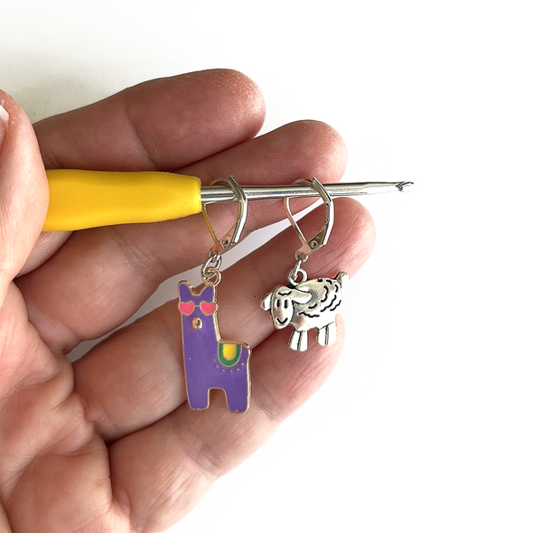 Purple enamel llama crochet stitch marker with pink heart eyes and yellow and green accents plus antique silver lamb locking crochet stitch markers on crochet hook held in a hand, made and sold by Pretty Warm Designs