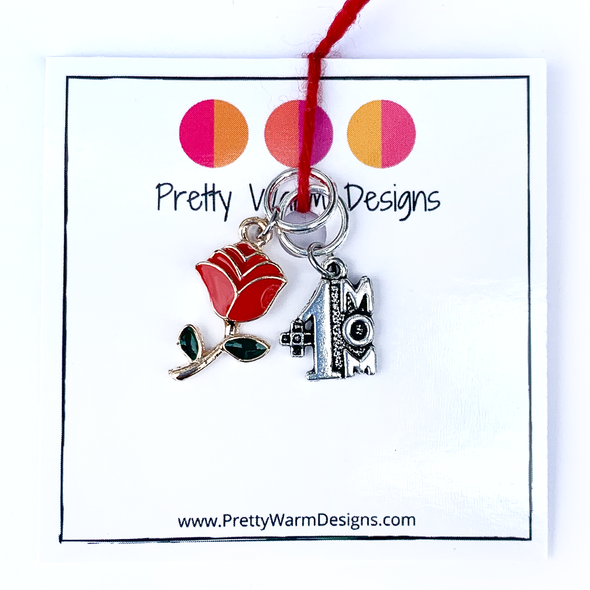 Two Mother's Day-themed knitting ring stitch markers, one red and green enamel rose and one silver #1 Mom attached with yarn to cardstock with Pretty Warm Designs text and logo