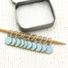 Set of ten blue on gold-toned number charm ring stitch markers on bamboo knitting needle with tin displayed on white knitted square by Pretty Warm Designs