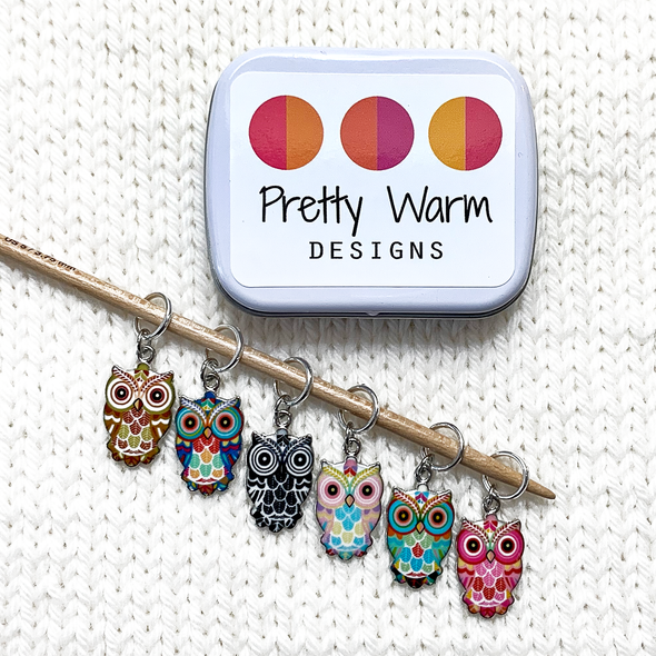 Set of six multi-coloured enamel owl charm snag free ring knitting stitch markers with decorative storage tin on knitting needle and white knitted background by Pretty Warm Designs