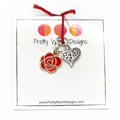 Two Valentine's Day themed knitting ring stitch markers, one red rose and one Tibetan antiqued silver filigree heart attached with red yarn to cardstock with Pretty Warm Designs text and logo