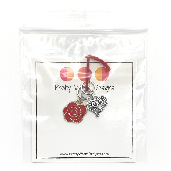 Two Valentine's Day themed knitting ring stitch markers, one red rose and one Tibetan antiqued silver filigree heart attached with red yarn to cardstock with Pretty Warm Designs text and logo in a poly bag