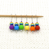 Set of six red, orange, yellow, green, blue and purple snag free silicone beads and rainbow glass beads on nylon coated wire, stitch markers on knitting needle placed on white knitted fabric by Pretty Warm Designs
