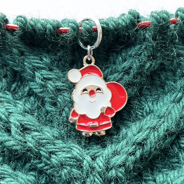 One red, white, black and gold Christmas Santa holding a toy sack knitting stitch marker threaded on a red knitting needle cable resting on a green knitted background and sold by Pretty Warm Designs Inc.