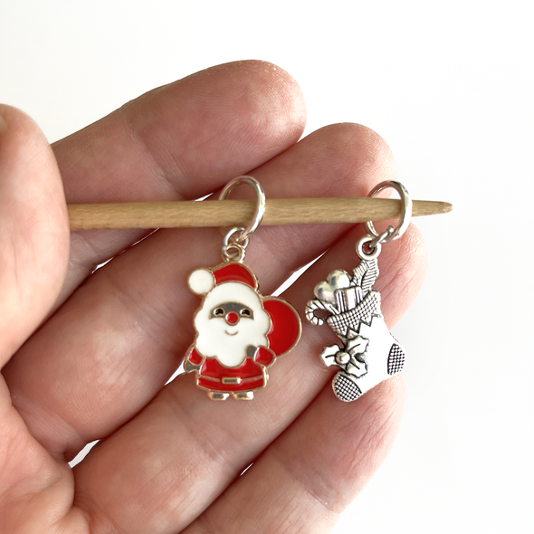 Two Christmas-themed knitting ring stitch markers, one red and white Santa with toy sack and one silver toned Christmas stocking held on bamboo knitting needle made by Pretty Warm Designs