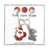 Two Christmas-themed knitting ring stitch markers, one red and white Santa with toy sack and one silver toned Christmas stocking attached to silver ring on cardstock printed with Pretty Warm Designs logo and URL
