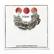 20 small heart stitch markers strung on a silver earring hoop attached to card stock with Pretty Warm Designs logo and text