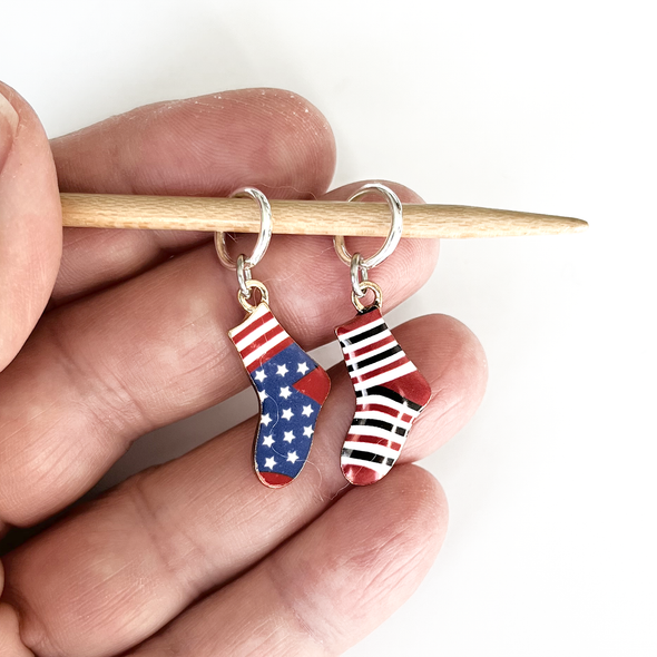 Two red, white and blue or black enamel socks knitting ring stitch markers on bamboo knitting needle held in a hand made and sold by Pretty Warm Designs