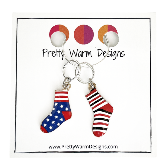 Two red, white and blue or black enamel socks knitting ring stitch markers attached with silver hoop ring to cardstock with Pretty Warm Designs text and logo