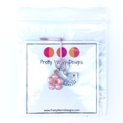 Two spring-themed knitting ring stitch markers, one pink enamel cherry blossom and one small silver song bird attached with yarn to cardstock in poly bag with Pretty Warm Designs text and logo