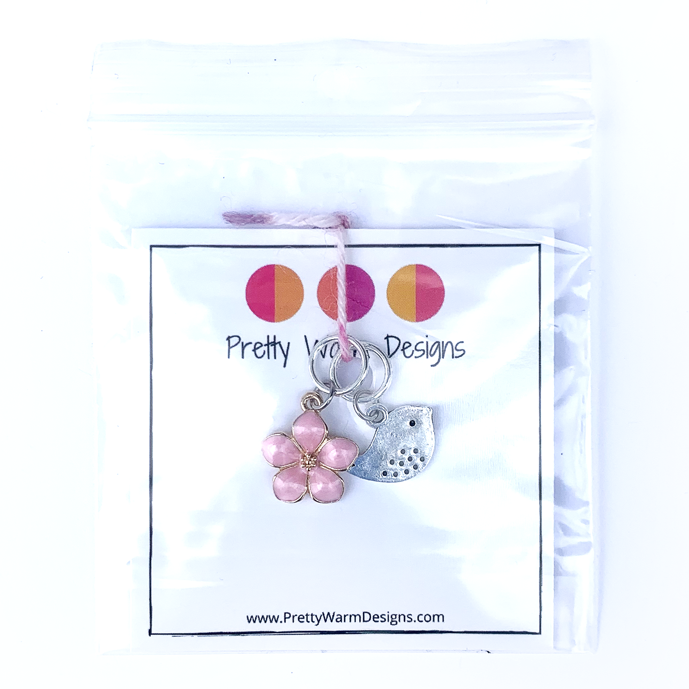 Two spring-themed knitting ring stitch markers, one pink enamel cherry blossom and one small silver song bird attached with yarn to cardstock in poly bag with Pretty Warm Designs text and logo