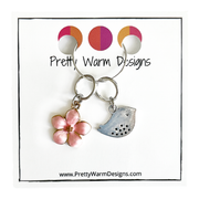 Two spring-themed knitting ring stitch markers, one pink enamel cherry blossom and one small silver song bird attached with silver hoop ring to cardstock with Pretty Warm Designs text and logo