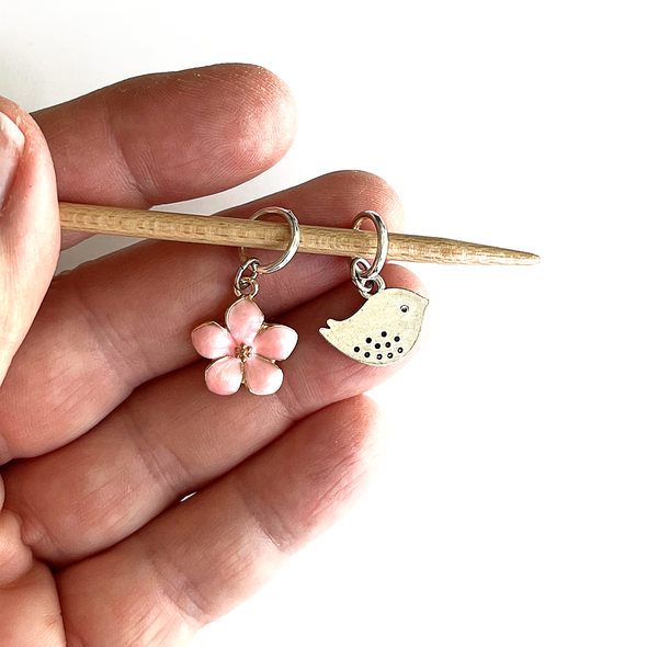Two spring-themed knitting ring stitch markers, one pink enamel cherry blossom and one small silver alloy song bird on bamboo knitting needle held in a hand, made and sold by Pretty Warm Designs