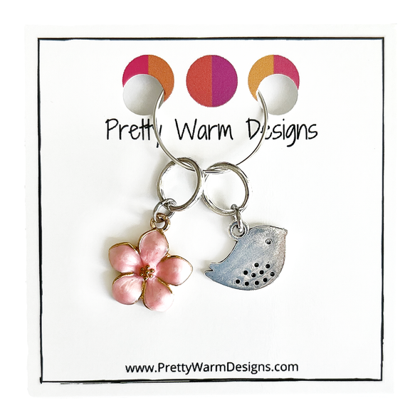 Two spring-themed knitting ring stitch markers, one pink enamel cherry blossom and one small silver song bird attached with silver hoop ring to cardstock with Pretty Warm Designs text and logo