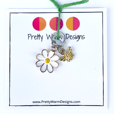 Two summer-themed knitting ring stitch markers, one yellow and white enamel daisy and one gold toned honeybee attached with yarn to cardstock with Pretty Warm Designs text and logo