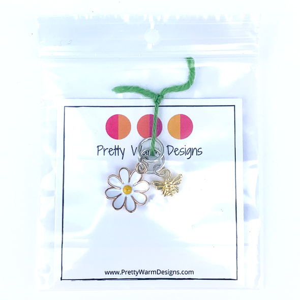 Two summer-themed knitting ring stitch markers, one yellow and white enamel daisy and one gold toned honeybee attached with green yarn to cardstock with Pretty Warm Designs text and logo in a poly bag