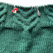 One red white and gold double toadstool knitting stitch marker and one silver alloy gnome knitting stitch marker threaded on a red knitting needle cable laying on a green knitting background sold by Pretty Warm Designs Inc.