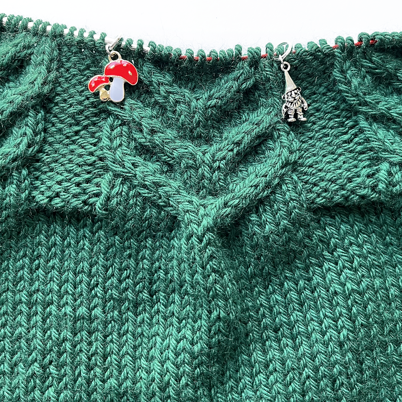 One red white and gold double toadstool knitting stitch marker and one silver alloy gnome knitting stitch marker threaded on a red knitting needle cable laying on a green knitting background sold by Pretty Warm Designs Inc.
