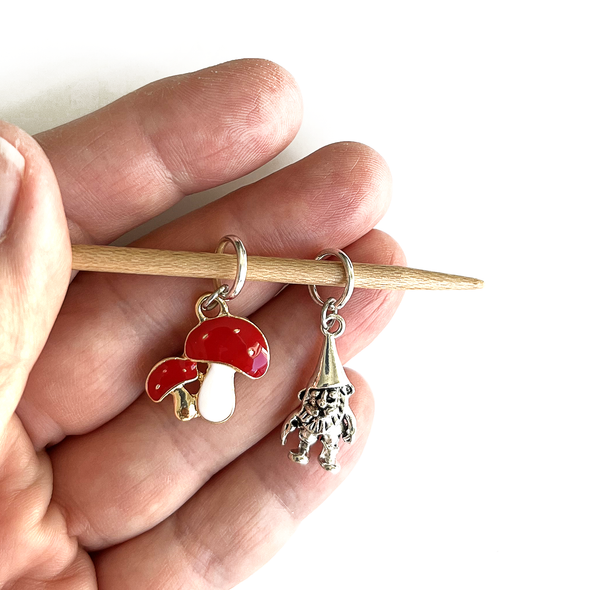 One red and white enamel toadstools and one silver alloy woodland gnome stitch markers for knitting held on bamboo knitting needle in a hand, made and sold by Pretty Warm Designs
