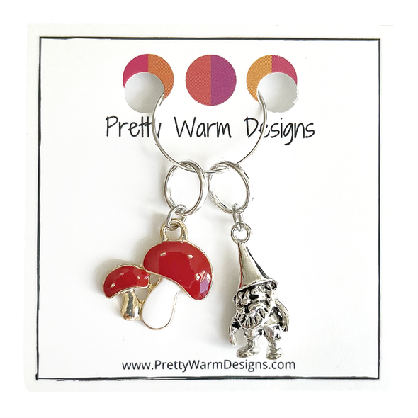 One red and white enamel toadstool knitting stitch marker and one antiqued silver gnome knitting stitch marker attached to silver ring hanging from white cardstock printed with Pretty Warm Designs logo and website URL