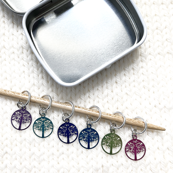 Six colour coated stainless steel round charm knitting stitch markers on bamboo needle sitting on white yarn knitted background with storage tin by Pretty Warm Designs Inc