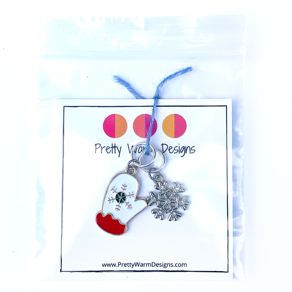 Two Winter-themed knitting ring stitch markers, one red, white and green enamel mitten and one silver toned snowflake with rhinestones attached with blue yarn to cardstock with Pretty Warm Designs text and logo in a poly bag
