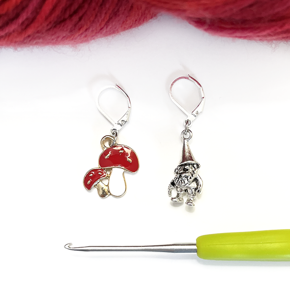 One red and white enamel toadstool crochet stitch marker and one antiqued silver gnome crochet stitch marker with red yarn and green handled crochet hook by Pretty Warm Designs Inc.