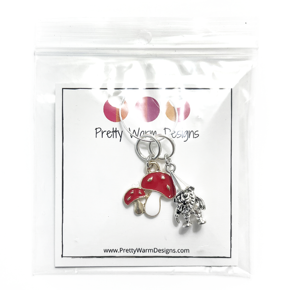 One red and white enamel toadstool knitting stitch marker and one antiqued silver gnome knitting stitch marker attached to silver ring hanging from white cardstock printed with Pretty Warm Designs logo and website URL packaged in clear poly bag