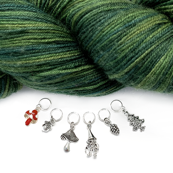 Set of six stitch markers for knitting, including a red enamel toadstool, as well as an antique silver acorn, toadstool, woodland gnome, pine cone and Christmas tree with skein of variegated green yarn sold by Pretty Warm Designs Inc.
