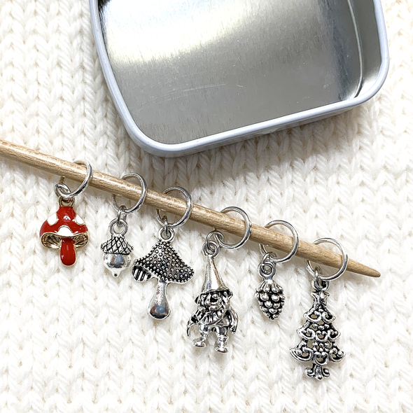 Set of six stitch markers for knitting, including a red enamel toadstool, as well as an antique silver acorn, toadstool, woodland gnome, pine cone and Christmas tree placed on bamboo knitting needle resting on a white knitted background with open tin sold by Pretty Warm Designs Inc.