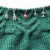 Five stitch markers for knitting, including a red enamel toadstool, as well as a toadstool, woodland gnome, pine cone and Christmas tree knitting stitch markers threaded on a red knitting needle cable resting on a green knitted background and sold by Pretty Warm Designs Inc.