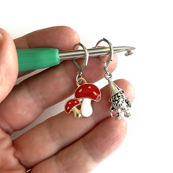 One red and white enamel toadstool crochet stitch marker and one antiqued silver gnome crochet stitch marker on crochet hook held in a hand, made and sold by Pretty Warm Designs